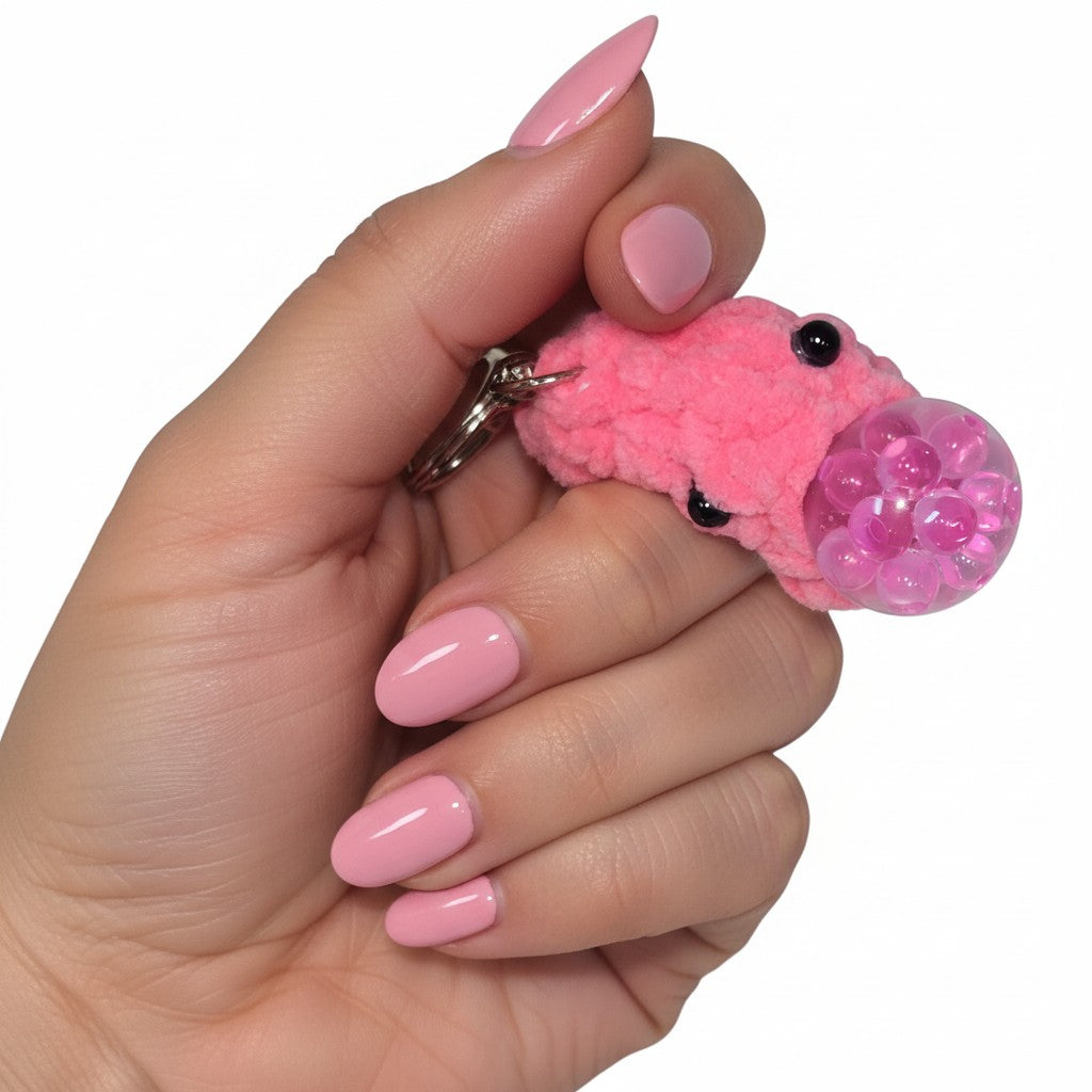 The Strawberry Shop pink crocheted blob stress ball keychain with bead eyes held in a hand with matching pink nails.